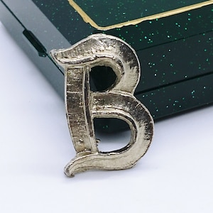 May include: A silver-toned metal brooch in the shape of the letter "B". The brooch has a textured surface and a pin on the back. The brooch is set against a dark green box with gold trim.