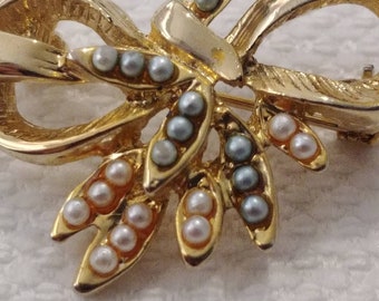 Puede incluir: Broche de tono dorado con un diseño de lazo y detalles de perlas. Las perlas son en tonos de blanco y azul.