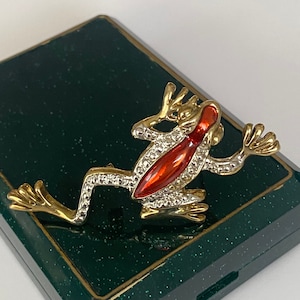 Może przedstawiać: Złota i czerwona broszka w kształcie żaby z cyrkoniami. Żaba siedzi na zielonym tle.