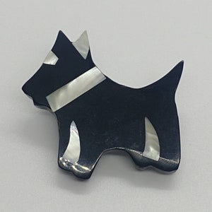 Może przedstawiać: Czarno-biała broszka w kształcie psa rasy Scottish Terrier. Broszka ma czarny korpus z białymi akcentami na kołnierzu, uszach i nogach. Pies jest pokazany z profilu.