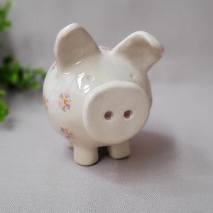 Può includere: Un piccolo salvadanaio in ceramica color crema chiaro, decorato con piccoli motivi floreali color lavanda. Il salvadanaio ha un corpo rotondo, quattro piccole zampe e un grugno prominente con due narici.