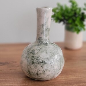 Puede incluir: Un jarrón de cerámica hecho a mano con una base bulbosa y un cuello delgado. El jarrón presenta una superficie moteada y texturizada con tonos de blanco, gris y verde. El jarrón mide aproximadamente 20 cm de altura y es adecuado para exhibir flores o como pieza decorativa.