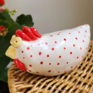 May include: A ceramic hen figurine with a white body and red polka dots. The hen has a red comb and wattle, and a yellow beak. The figurine is sitting on a woven wicker basket.