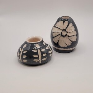 Può includere: Due piccoli vasi in ceramica con disegni floreali bianchi su sfondo nero. Il vaso a sinistra ha un motivo ripetitivo di foglie e punti. Il vaso a destra ha un unico grande fiore.