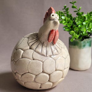 Könnte beinhalten: Handgefertigte Keramik-Hühnerfigur mit weißem Körper und rotem Kamm und Kehllappen. Das Huhn hat einen strukturierten, abgerundeten Körper und ein detailliertes Gesicht. Die Figur ist ein dekorativer Artikel.