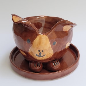 May include: A brown and tan ceramic planter shaped like a dog with black painted eyes, nose, and mouth. The planter has a saucer.