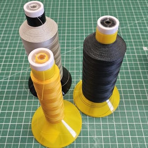 Pu&ograve; includere: Tre grandi rocche di filo nei colori giallo, nero e grigio chiaro, ciascuna dotata di una base conica in plastica e un tappo bianco. Le rocche sono disposte su un tappetino da taglio verde con un righello visibile che mostra le misure in centimetri.