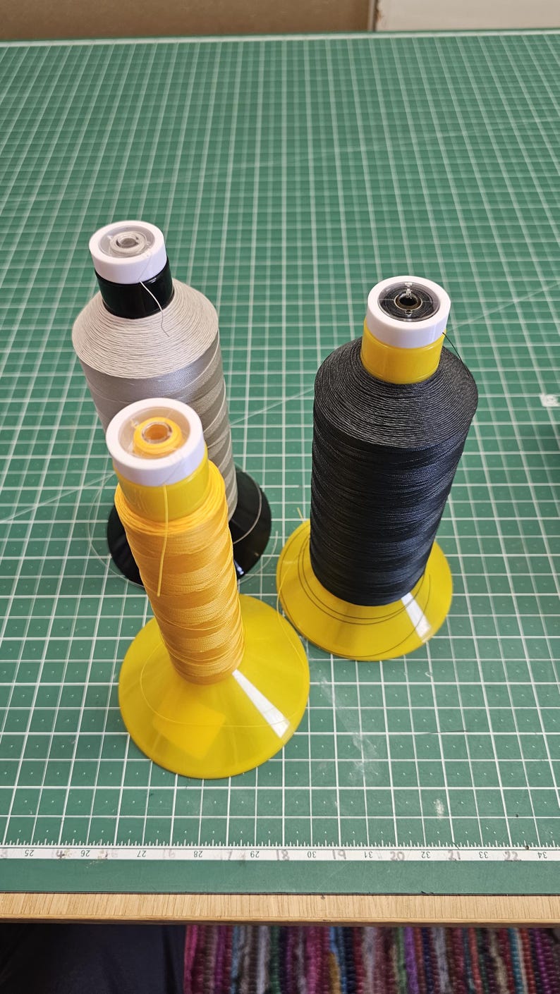 Pu&ograve; includere: Tre grandi rocche di filo nei colori giallo, nero e grigio chiaro, ciascuna dotata di una base conica in plastica e un tappo bianco. Le rocche sono disposte su un tappetino da taglio verde con un righello visibile che mostra le misure in centimetri.