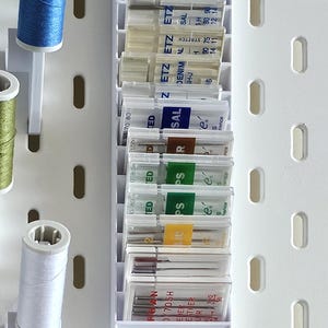Può includere: Un organizzatore in plastica bianca con scomparti trasparenti contiene diversi aghi da cucito. Gli aghi sono etichettati con diverse dimensioni e tipi, tra cui "Denim", "Stretch", "Sal", "Ps", "R" e "Ted".