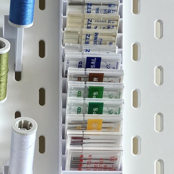 May include: A white plastic organizer with clear compartments holds various sewing needles. The needles are labeled with different sizes and types, including "Denim", "Stretch", "Sal", "Ps", "R", and "Ted".