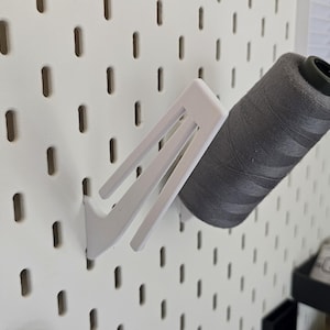 May include: A white pegboard with a white thread spool holder holding a grey spool of thread. The pegboard has evenly spaced holes. The thread spool is cylindrical and has a black top. The holder is angled.