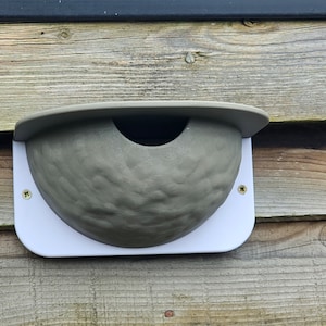 May include: A grey birdhouse with a white trim, mounted on a wooden wall. The birdhouse has a rounded shape and a small opening for birds to enter.