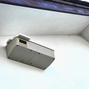 May include: A rectangular, light gray plastic housing unit mounted to a white exterior wall. The device features a small dark rectangular sensor window on the front, a series of horizontal ventilation slats, and several small circular screw holes.