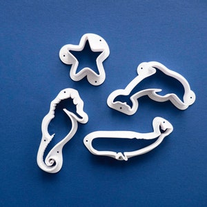 May include: Four white plastic cookie cutters shaped like a starfish, a dolphin, a whale, and a seahorse. The cutters are on a blue background.