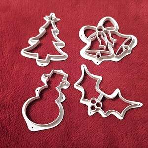 May include: Four white plastic cookie cutters shaped like a Christmas tree, a snowman, a holly leaf, and a bell with a bow.