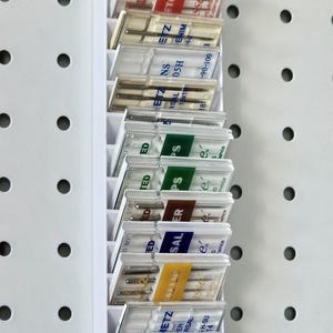 Könnte beinhalten: Ein weißer Kunststoff-Organizer mit mehreren transparenten Kunststoffboxen für Nähmaschinennadeln. Die Boxen haben Etiketten mit verschiedenen Farben und Texten, darunter "ETCH", "ENIM", "PS" und "SALER". Der Organizer ist an einer weißen Lochwand befestigt.