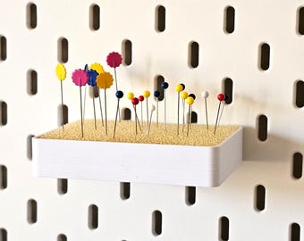 May include: A white pegboard with a rectangular white shelf holding various colorful pins. The pins have decorative tops in yellow, pink, blue, and red. The pegboard has evenly spaced, vertical oval holes.