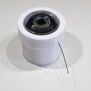 Pu&ograve; includere: Un portabobina cilindrico in plastica bianca contenente una rocchetto di filo nero. Il supporto presenta un coperchio circolare trasparente con una piccola apertura, che permette a un singolo filo nero di uscire dal lato dell'involucro bianco.