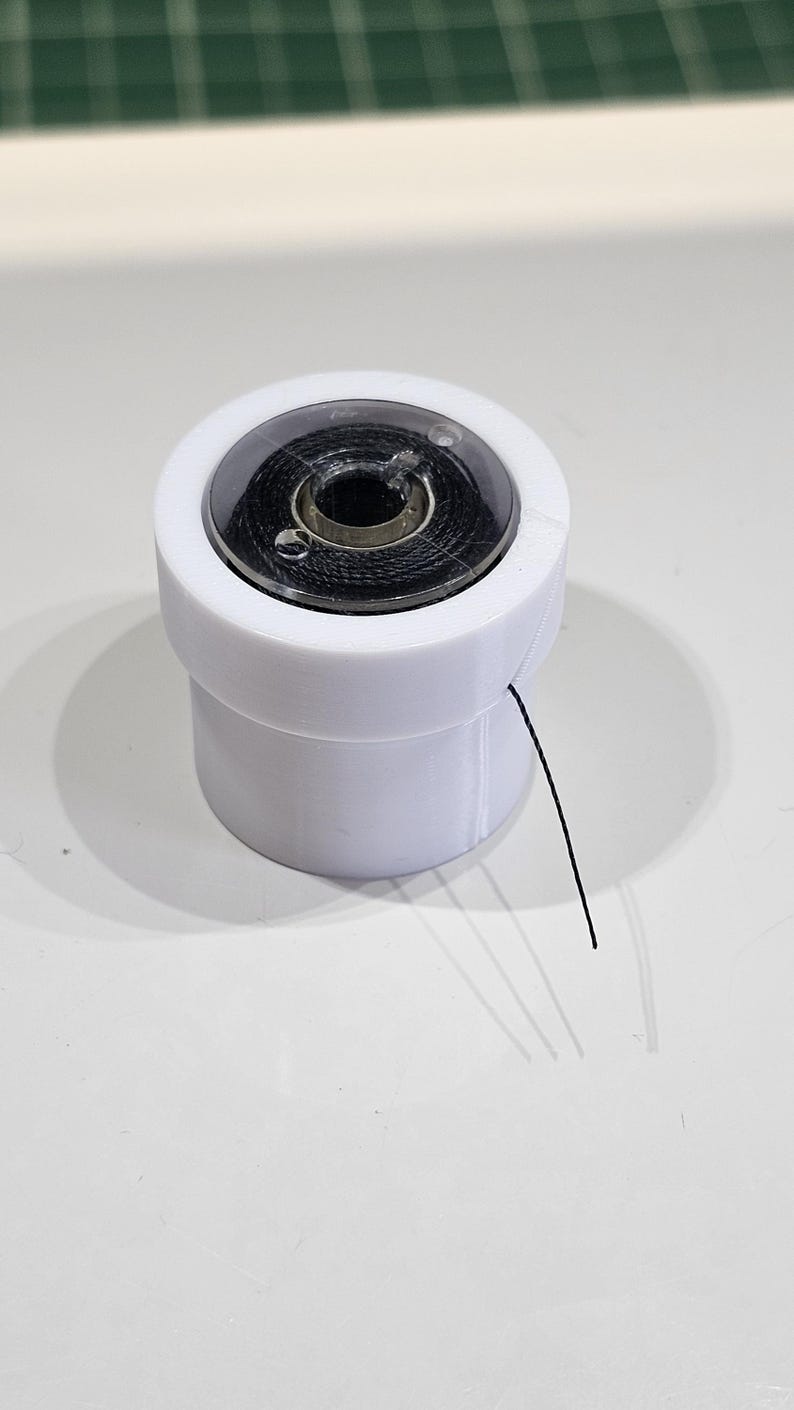 Pu&ograve; includere: Un portabobina cilindrico in plastica bianca contenente una rocchetto di filo nero. Il supporto presenta un coperchio circolare trasparente con una piccola apertura, che permette a un singolo filo nero di uscire dal lato dell'involucro bianco.