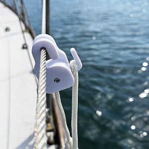 May include: A white plastic boat rope hook attached to a metal cable. The hook is designed to hold a rope and is secured with a screw. A white rope is also attached. The background shows the sea, out of focus.