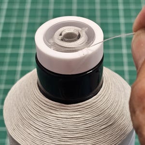 May include: A large spool of white sewing thread featuring a black plastic neck and a white top cap. A thin, translucent thread is being pulled from the center of the spool, which is wound with tightly coiled white thread in a conical shape.