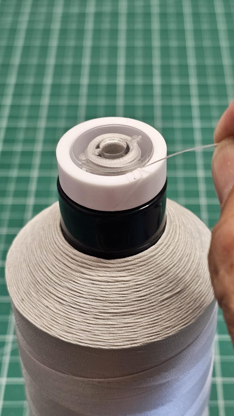 Pu&ograve; includere: Una grande rocca di filo da cucito bianco con un collo in plastica nera e un tappo superiore bianco. Un filo sottile e traslucido viene estratto dal centro della rocca, avvolta con filo bianco ben stretto in una forma conica.
