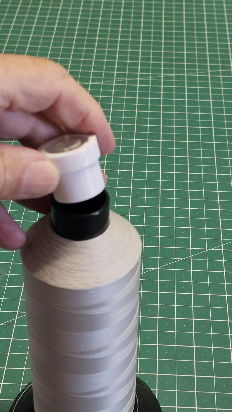 Pu&ograve; includere: Una grande bobina di filo grigio chiaro con un nucleo in plastica nera e un tappo in plastica bianca rimovibile. Il filo &egrave; avvolto strettamente attorno alla bobina cilindrica, posizionata in verticale per mostrare la trama del materiale.