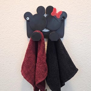 Könnte beinhalten: Ein schwarzer Wand-Handtuchhalter mit zwei kreisförmigen Silhouetten, die an Mausköpfe erinnern, wobei einer mit einer roten Schleife verziert ist. Zwei runde Haken ragen aus der Basis hervor und halten ein weinrotes und ein schwarzes Handtuch.