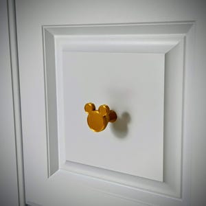 Op de afbeelding: Een goudkleurige kastknop in de vorm van een muizenkop is bevestigd op een witte kastdeur. De knop heeft een cirkelvormig lichaam met twee kleinere cirkels erboven, die de oren vormen.
