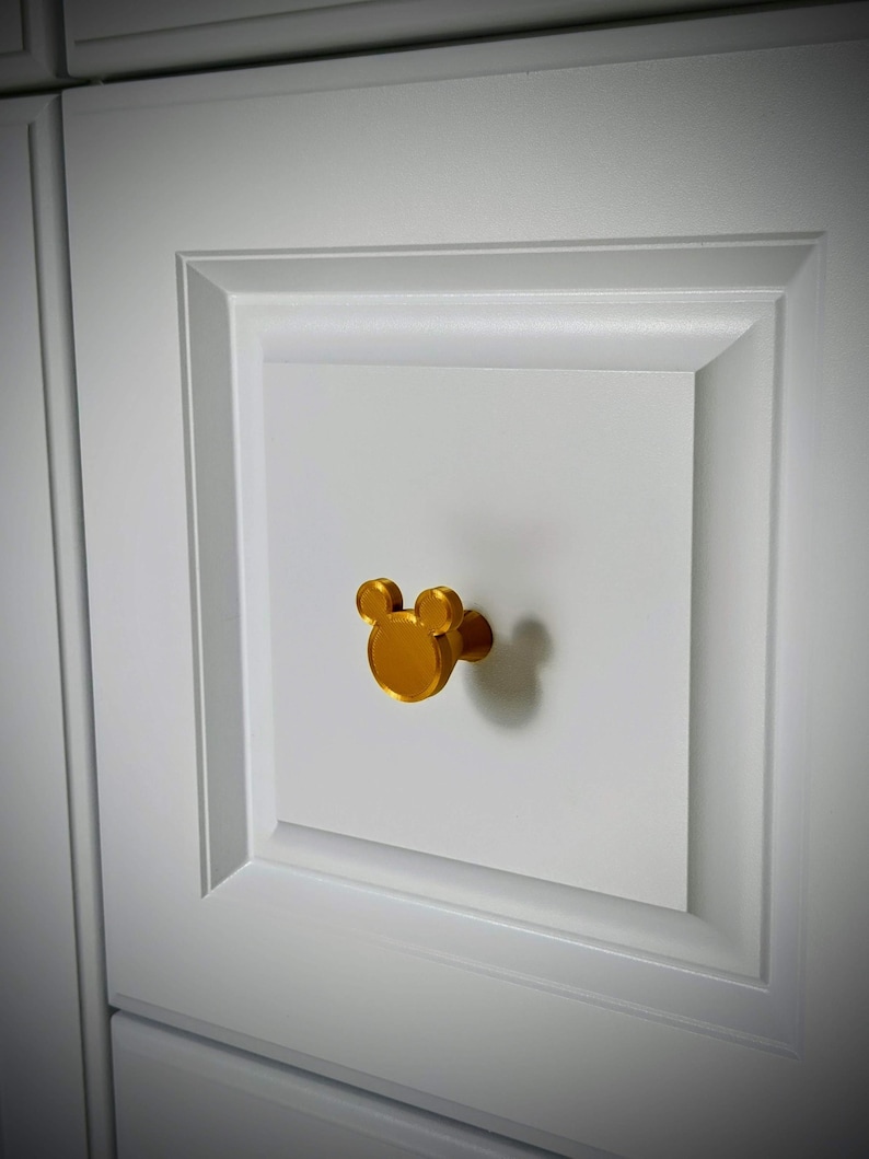 Op de afbeelding: Een goudkleurige kastknop in de vorm van een muizenkop is bevestigd op een witte kastdeur. De knop heeft een cirkelvormig lichaam met twee kleinere cirkels erboven, die de oren vormen.