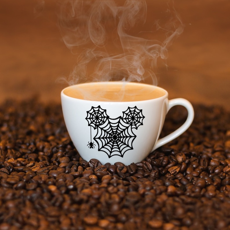 May include: A white coffee mug with a black spiderweb design in the shape of Mickey Mouse ears. The mug is filled with coffee and steam is rising from the top. The mug is sitting on a bed of coffee beans.