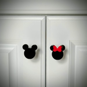 Op de afbeelding: Twee zwarte kastknoppen in de vorm van Mickey Mouse-hoofden, &eacute;&eacute;n met een rode strik. De knoppen zijn gemonteerd op witte kastdeuren. Deze knoppen zijn een leuke en grillige toevoeging aan elke kamer.