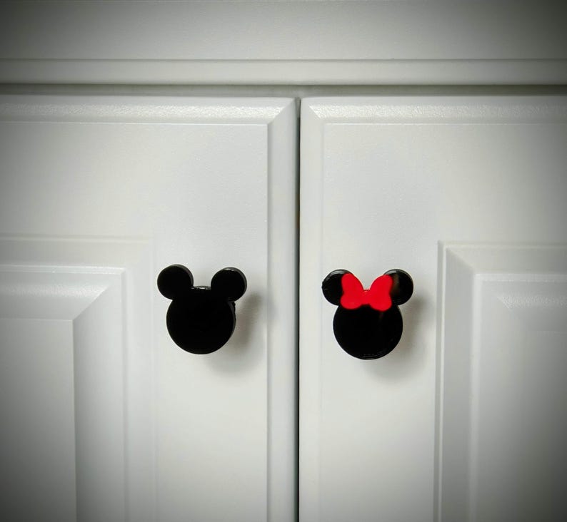 Op de afbeelding: Twee zwarte kastknoppen in de vorm van Mickey Mouse-hoofden, &eacute;&eacute;n met een rode strik. De knoppen zijn gemonteerd op witte kastdeuren. Deze knoppen zijn een leuke en grillige toevoeging aan elke kamer.