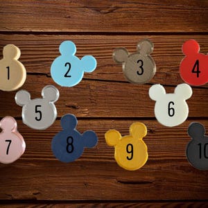 Op de afbeelding: Een verzameling van tien Mickey Mouse-vormige items in verschillende kleuren, waaronder beige, blauw, bruin, rood, zilver, wit, roze, marine, geel en zwart. Elk item is genummerd van 1 tot 10, op een donkere houten achtergrond.