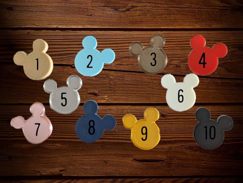 Op de afbeelding: Een verzameling van tien Mickey Mouse-vormige items in verschillende kleuren, waaronder beige, blauw, bruin, rood, zilver, wit, roze, marine, geel en zwart. Elk item is genummerd van 1 tot 10, op een donkere houten achtergrond.