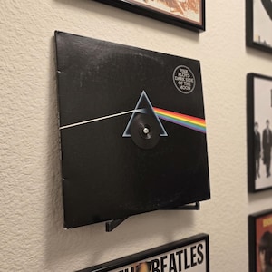 May include: A black vinyl record sleeve for Pink Floyd's Dark Side of the Moon album, featuring a white light beam entering a prism and exiting as a rainbow spectrum. The sleeve is mounted on a wall-mounted display stand with a central screw.