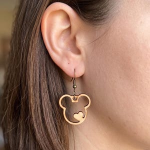 May include: Wooden earrings featuring a Mickey Mouse head design with a heart cutout. The earrings are light brown and hang from bronze-colored hooks. The earrings are a fun accessory for Disney fans.