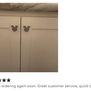 Op de afbeelding: Twee witte kastdeuren met zilverkleurige Mickey Mouse-vormige knoppen. De knoppen zijn gecentreerd op de deuren, die gesloten zijn. Op de achtergrond is een lichtgekleurd aanrechtblad te zien.