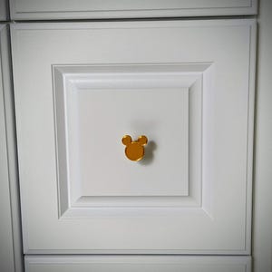 Op de afbeelding: Een goudkleurige kastknop in de vorm van een muizenkop is gecentreerd op een witte kastdeur. De knop heeft een glad, reflecterend oppervlak. De kastdeur maakt deel uit van een set witte kasten.