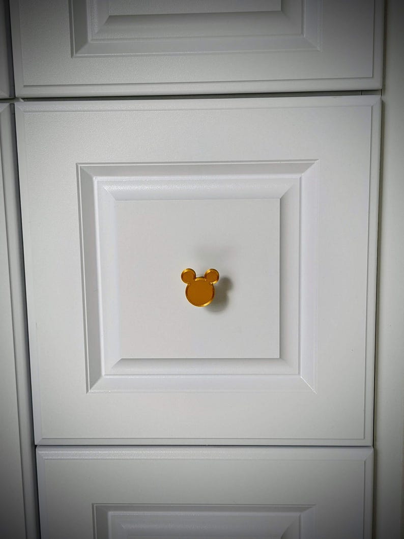 Op de afbeelding: Een goudkleurige kastknop in de vorm van een muizenkop is gecentreerd op een witte kastdeur. De knop heeft een glad, reflecterend oppervlak. De kastdeur maakt deel uit van een set witte kasten.