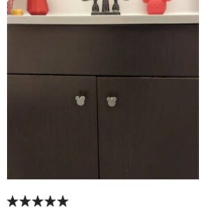 Op de afbeelding: Donkerbruine badkamerkast met twee deuren en zilveren Mickey Mouse-vormige knoppen. De kast staat onder een wit aanrechtblad met een zwarte kraan en een rode Mickey Mouse-vormige zeepdispenser.