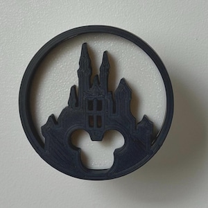 May include: A dark blue circular emblem featuring a castle silhouette within a circle. The castle design includes turrets and windows, with a Mickey Mouse head shape at the bottom. The emblem is likely a decorative item or a charm.