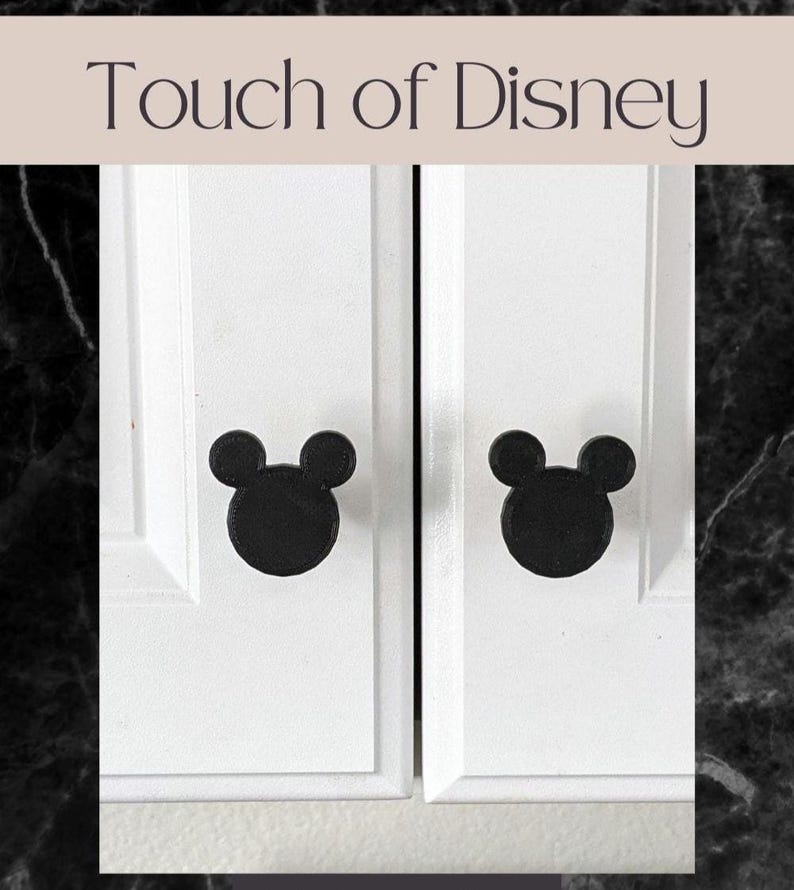 Op de afbeelding: Witte kastdeuren met zwarte Mickey Mouse-vormige knoppen. De tekst "Touch of Disney" wordt boven de deuren weergegeven. De knoppen geven de kast een vleugje vrolijkheid.