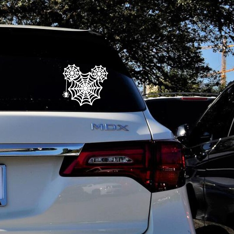 May include: White vinyl decal of a spider web shaped like Mickey Mouse's head with a small spider hanging from the web. The decal is on the back window of a white SUV.