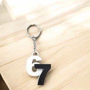 May include: A white and black number 67 keyring. The number 6 is white, and the number 7 is black. The keyring is attached to a silver metal ring and chain. The keyring is on a light brown wooden surface.