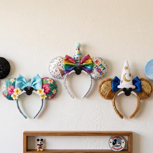 Pode incluir: Cinco tiaras decorativas com orelhas expostas numa parede. A coleção inclui uma tiara com laço preto de bolinhas, uma tiara de flores tropicais, uma tiara de aniversário com chapéu, uma tiara de mago e uma tiara de cor azul claro.