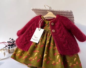 May include: A doll's outfit featuring a burgundy knit cardigan and a green dress with a leaf pattern. The dress has a round neckline and is buttoned in the front. The outfit is displayed on a wooden hanger.