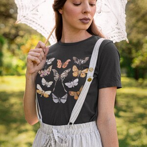 May include: A black t-shirt featuring a collection of butterfly illustrations in various colors. The person is wearing a striped skirt with suspenders and holding a white lace parasol. The background is a blurred green outdoor scene.