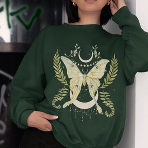 May include: A dark green sweatshirt with a white and green moth design. The moth is surrounded by leaves and a crescent moon. The sweatshirt has a crew neck and long sleeves.