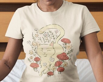 Puede incluir: Una mujer lleva una camiseta de color crema con un gato de dibujos animados rodeado de setas y flores. Está sentada en una cama con un gato en su regazo.